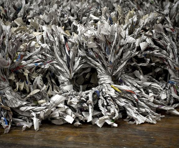 Newspapers are folded into thin tubes and tied together. The many tubes tied together look like brushes made from newspaper. They are black and white with some pops of color in between.