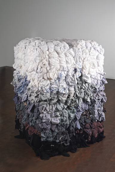 A square sculpture is standing on the floor and its surface is covered in textiles, possibly shirts, dyed in different colors. On top, the textiles are white, and they slowly fade into a lilac and blue hue. The change of color has an ombre-effect. 