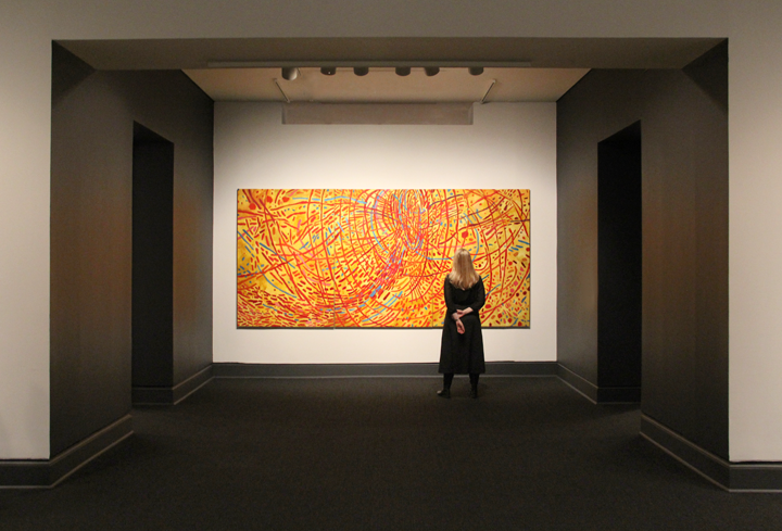 A woman with long, blonde hair is standing in a gallery looking at a large painting. Abstract painting features a vivid yellow background covered by circles, daubs, and straight and wavy lines in red, orange, cobalt, sky blue, and violet. Arcing red strokes evoke concentric circles. Straight lines in other hues radiate out from the center circle like a starburst.
