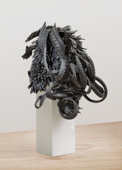 A sculpture made from black rubber tires sits on a pedestal in a room with a white wall and wooden floors. The black rubber tires are shaped and distorted in way that they create an organic form, resembling an insect or an alien-like figure.