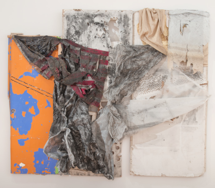 A mixed media piece consisting of cloth and painted wood hangs on a white wall. The cloth Is very distressed: Many holes are cut into the gray fabric and looks like something picked up from a dirty street. The cloths are draped over pieces of distressed canvas and wood painted in white and a bright orange, blue, and red. The red stripes resemble the stripes of the American flag.