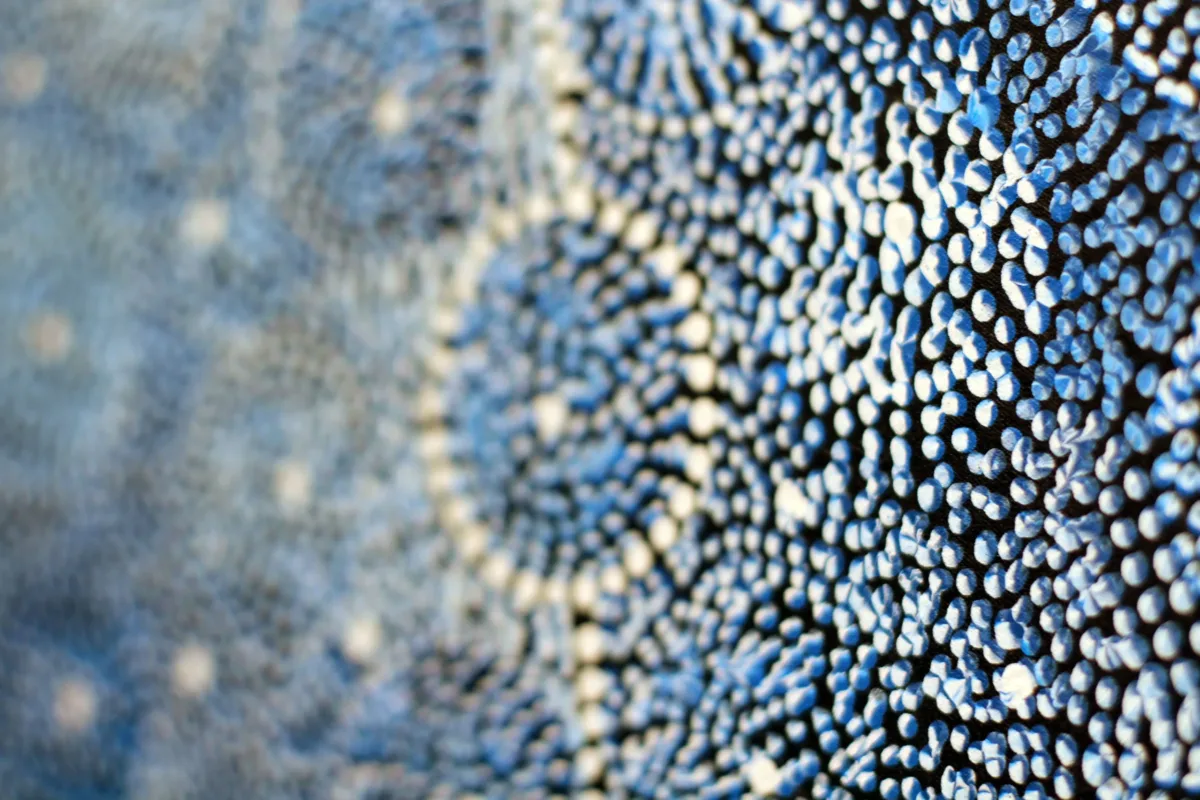 A very close-up detail shot of a painting featuring tiny dots arranged to form vary shades of blue circles outlined in white. Layered blue dots, appearing almost three-dimensional against a black background, are in focus in the foreground and the blue background is blurred.