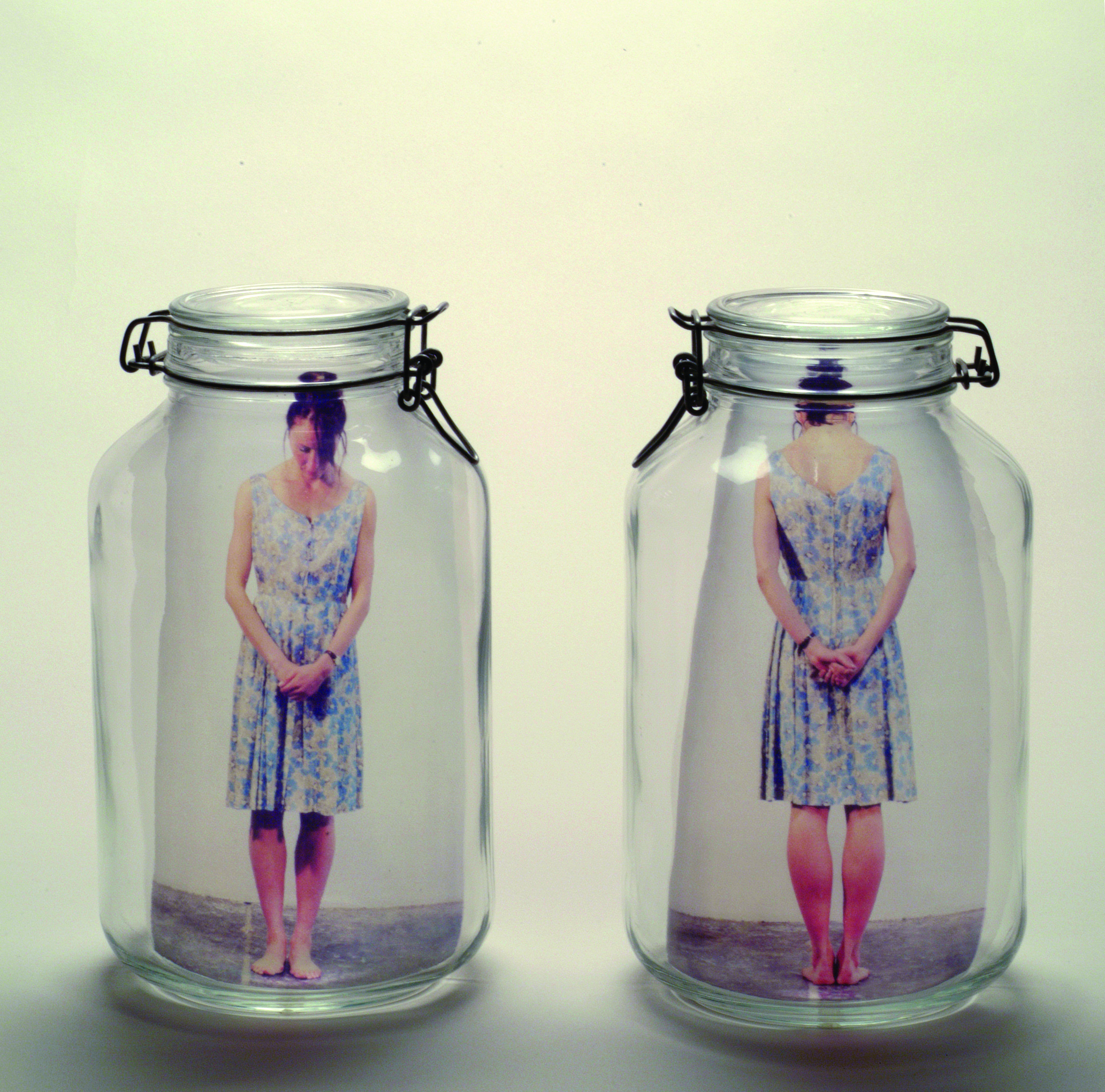 Two clear glass mason jars side by side. In the left jar is a color photograph of a light skinned woman standing with head bent and hands clasped in front of her. In the right jar is the same woman seen from rear with hands clasped behind her.