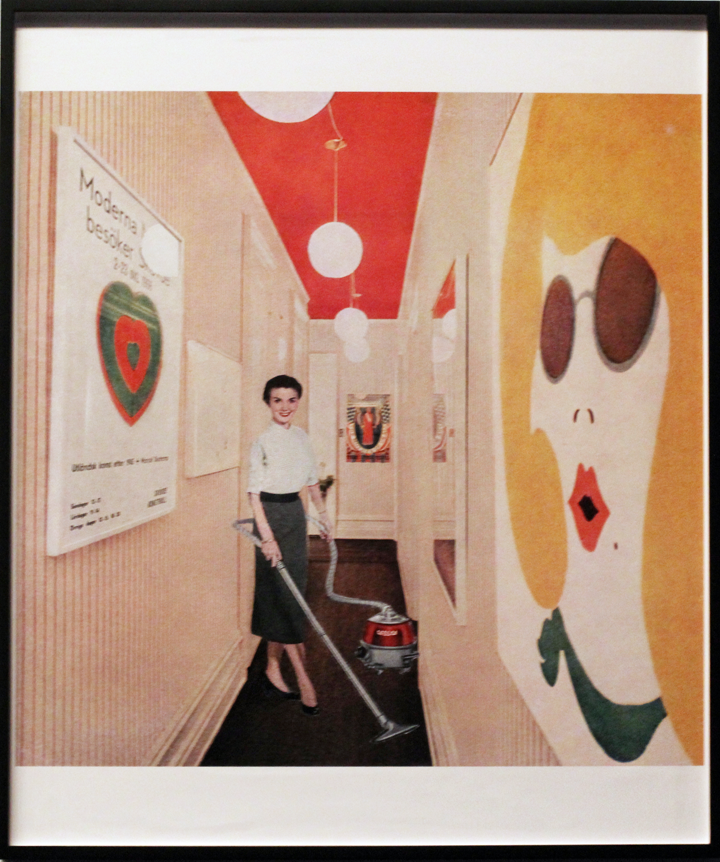 A light-skinned woman with dark hair poses with a vacuum in a hallway with a red ceiling. She wears a white, short-sleeved blouse, a grey pencil skirt, and black shoes. On the walls of the hallway are colorful framed images, including a large illustration of a blonde woman wearing sunglasses and green neck scarf. On the other side of the hall hangs a print of a green and red heart with indistinguishable text.