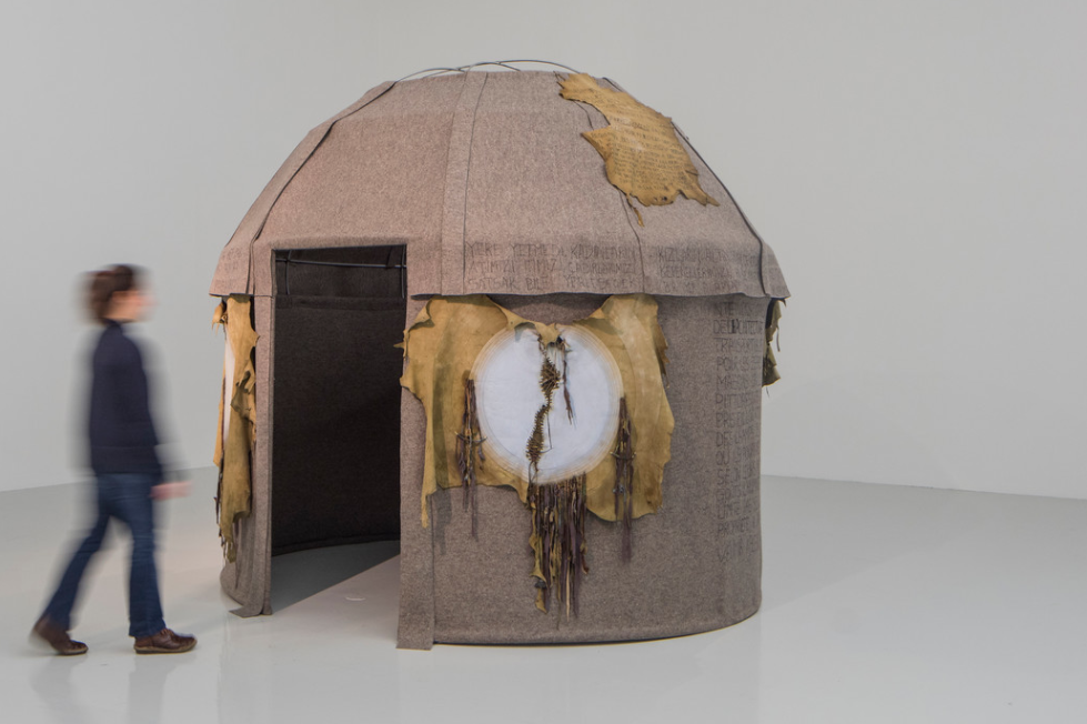 A life-size, circular hut made of felt stands in the center of a white gallery space. A light-skinned person (who is blurred in motion) walks towards thee rectangular entrance to the hut. The hut has a domed roof with animal skin fastened over circular, opaque windows and on the roof.