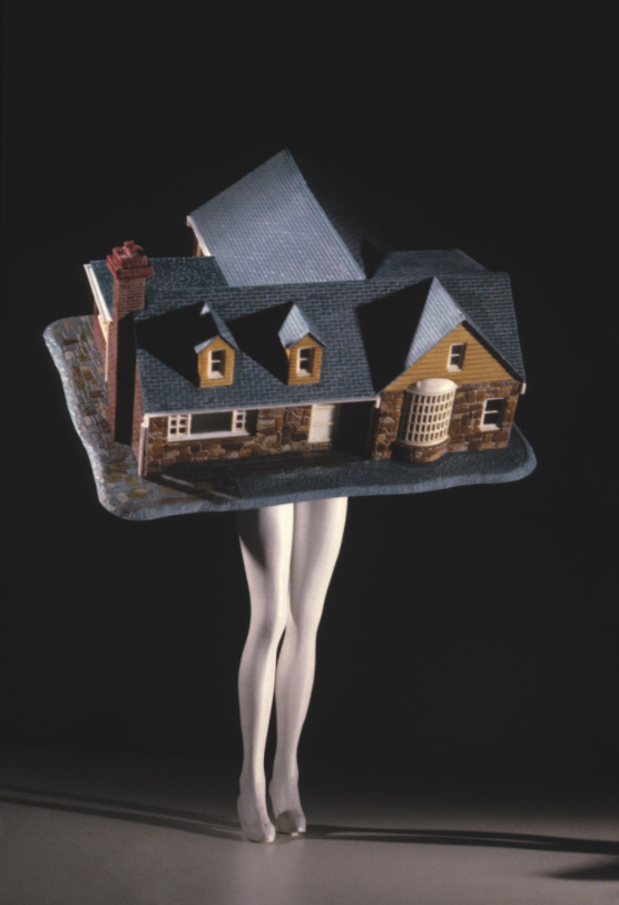 A photograph of a  pair of legs that resemble those of a store mannequin protrude from beneath a model of a red-brick house. The photograph's composition creates the impression that a woman is wearing or carrying the house.