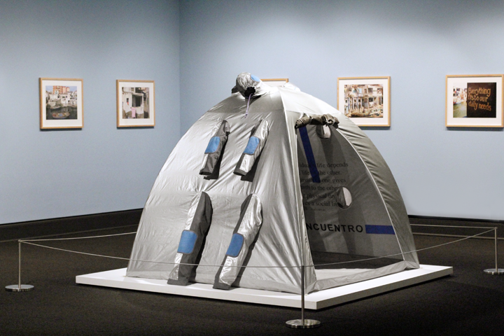 A grey camping tent sits in the center of a gallery space. It is roped off to prevent visitors from entering. The tent is dome-shaped with silver and blue tubular shapes attached to its exterior. Photographs of buildings hang in light-wood frames on grey walls behind the tent.