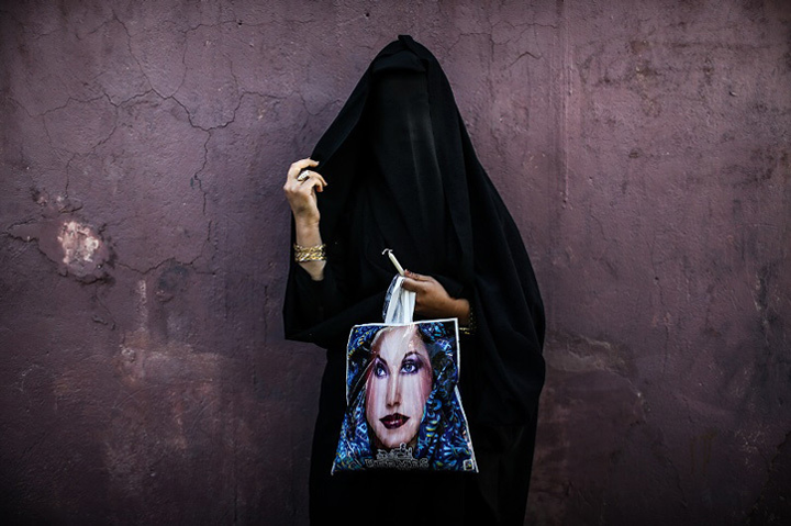 A photograph of a woman in a long, black burka stands before a concrete wall. In one hand, she holds her veil  and in the other, she holds a plastic bag with a woman's face printed on it.