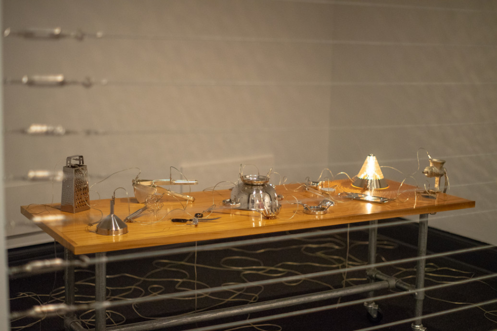 A rectangular table with a wooden surface stands in an empty room roped off by wires. On the table are various household objects like a cheese grater and a colander. The objects are connected by wires that spill out onto the floor. 