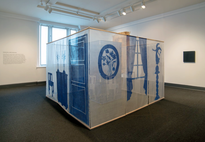 A large square structure made of transparent mesh material sits in the center of a gallery space. The exterior of the structure features blue silhouette illustrations of domestic objects like a dining table and window curtains. Inside the structure is a smaller mesh square that is blue with white silhouette illustrations of dining chairs.