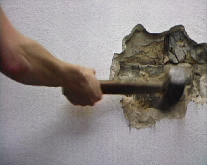 The arm of an unseen person swings a hammer into a white wall. The hammer has made a hole in the wall that reveals its concrete structure beneath the white paint.