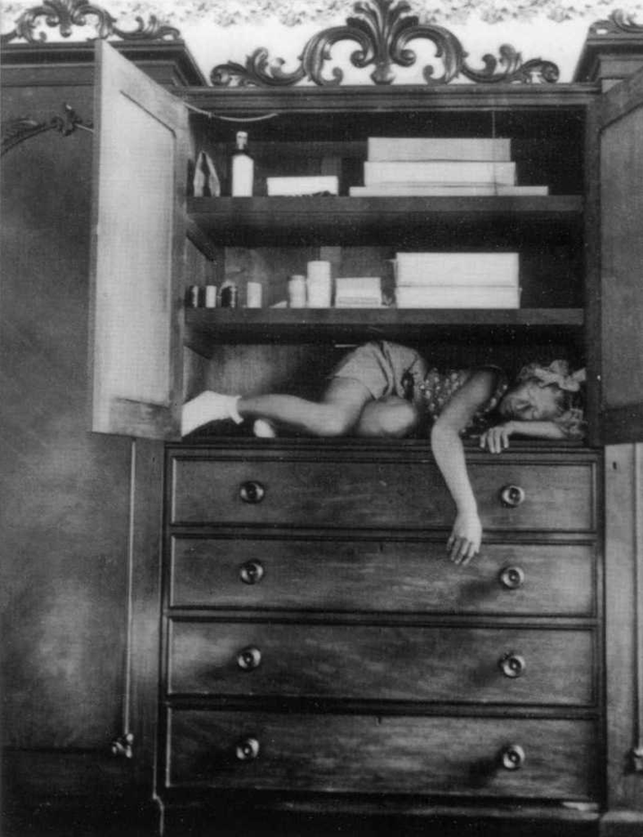 A black-and-white photograph of a light-skinned, light-haired girl sleeping on shelf of a large wooden cabinet. The cabinet doors are open to reveal the sleeping girl, who wears socks, shorts, a patterned top, and a hair bow. On the shelves above her are various boxes and canisters. The bottom half of the cabinet below the girl are four drawers that are closed.