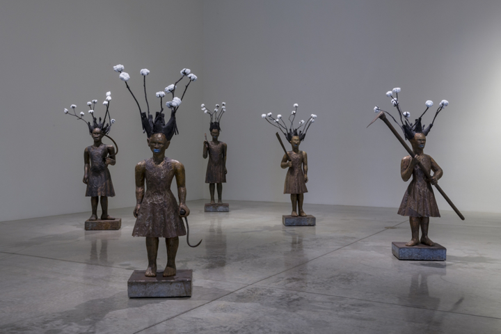 Five bronze sculptures of people standing apart from each other in a room on a shiny concrete floor. The figures are holding instruments to cut cotton, and from each of their heads, cotton balls are emerging on branches, as if there was cotton growing out of their heads.
