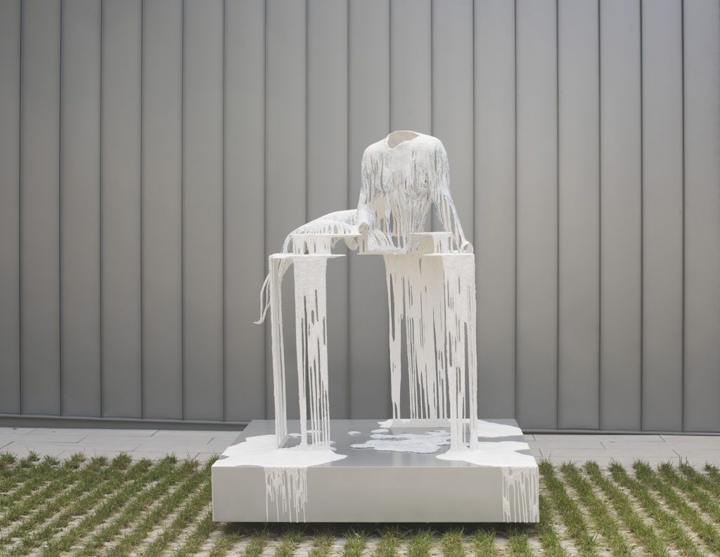 A white sculpture stands before a gray wall on a concrete ground that is covered in patches of grass. The sculpture is of a woman's body without a head sitting on a pedestal made from the same material. The material looks like paint dripping down, however, it has a solid form. 