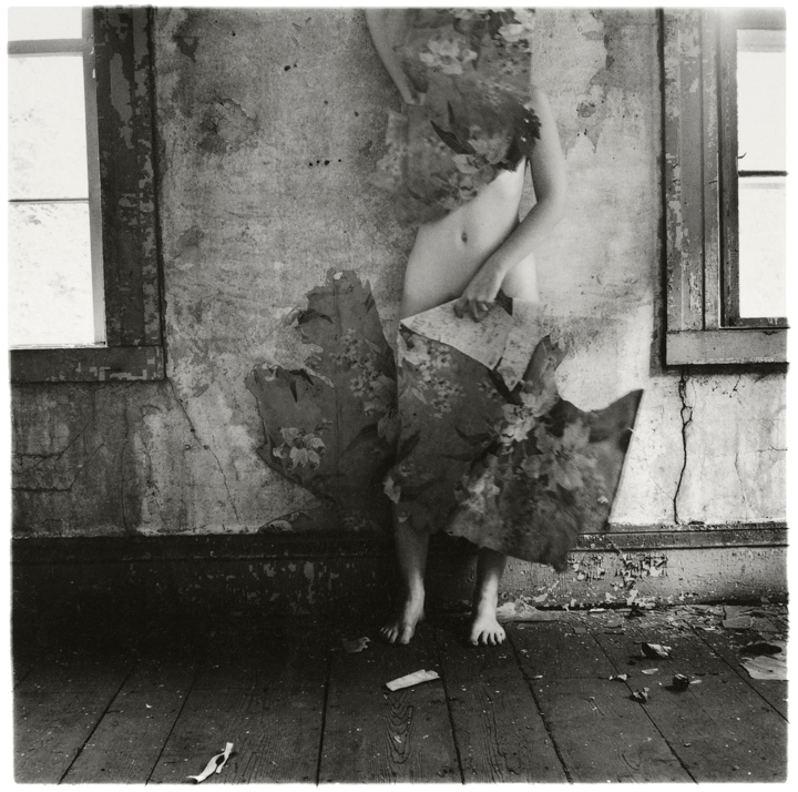 A black-and-white photograph of a nude woman stands in the interior of a run-down building. She poses between two dirty windows with chipped paint. The wall behind her is cracked and discolored.she holds pieces of floral wallpaper over her face and groin so only her stomach, arms, and feet are visible.