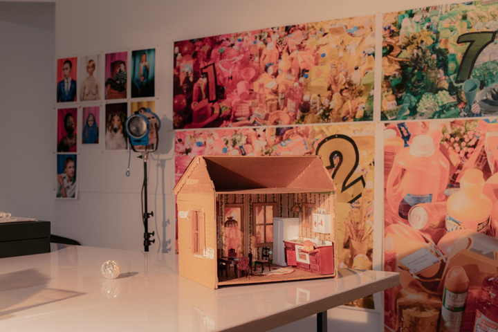 A photograph of a sun-filled interior scene. A dollhouse sits prominently on a white table. In the background, large photographs of plastic bottles hang on the white wall.