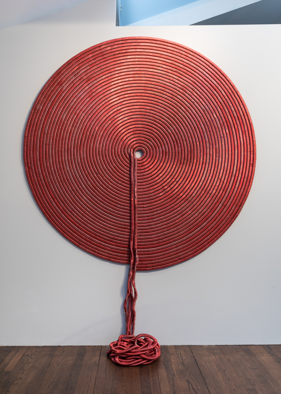 A large, circular wall sculpture hangs on a plain white wall. This red-colored sculpture resembles wrapped yarn, with the loose ends spilling from the center of the sculpture to the floor.