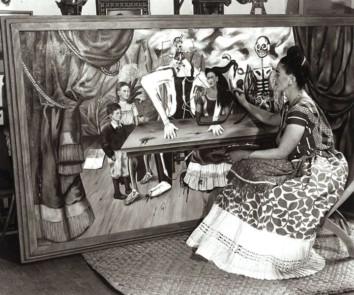 A black and white photo of a woman painting a large canvas in a wooden frame. The woman has a light skin tone, black hair in a bun, and wears a colorful, long dress with floral prints. The painting is of a group of people and abstract figures sat by a table. The person in the middle resembles the woman's appearance.