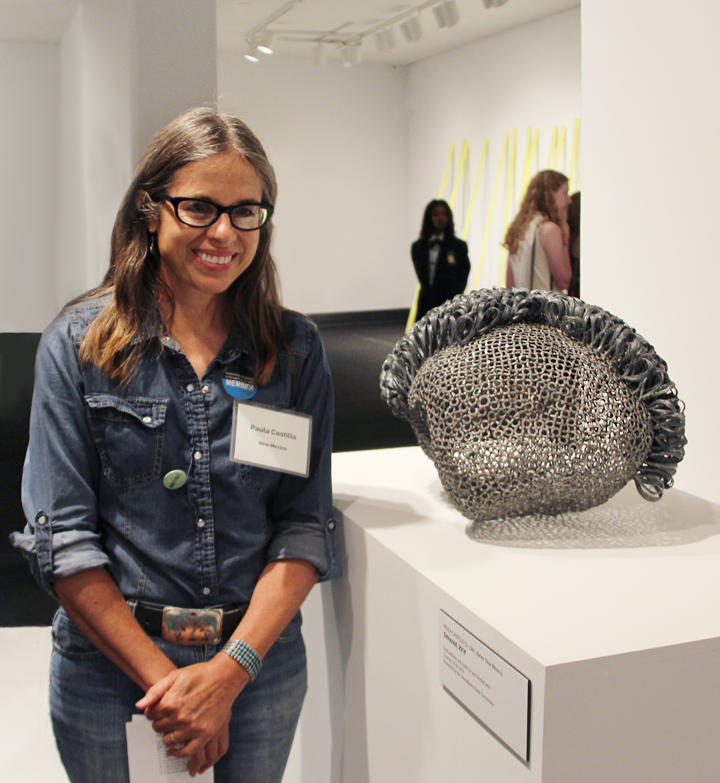 A woman with medium-light skin and brown with greying roots stands beside an abstract metal sculpture resembling mesh or chainmail. She wears dark-rimmed glasses, a denim button-down shirt, jeans, and a brown belt with a large silver buckle. She wears a name-tag that says “Paula Castillo.”