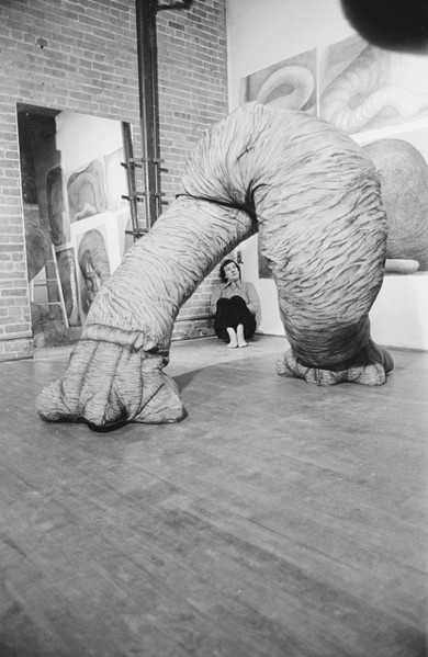 A black-and-white photograph of an artist's studio. At the center of the room is a large sculpture that seems to resemble the legs of an elephant. A light-skinned woman with short, dark hair sits on the floor in the corner of the room and looks at the sculpture. She wears a collared shirt, dark pants, and no shoes.
