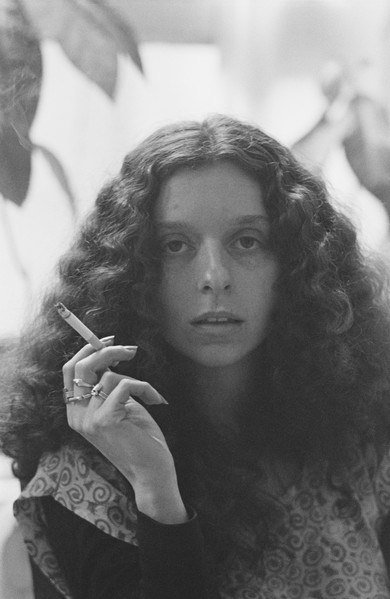 A black-and-white portrait of a light-skinned woman with long, curly hair. She stares directly at the camera and holds a cigarette in her right hand, on which she wears several rings. Her blouse is decorated with swirling geometric shapes.
