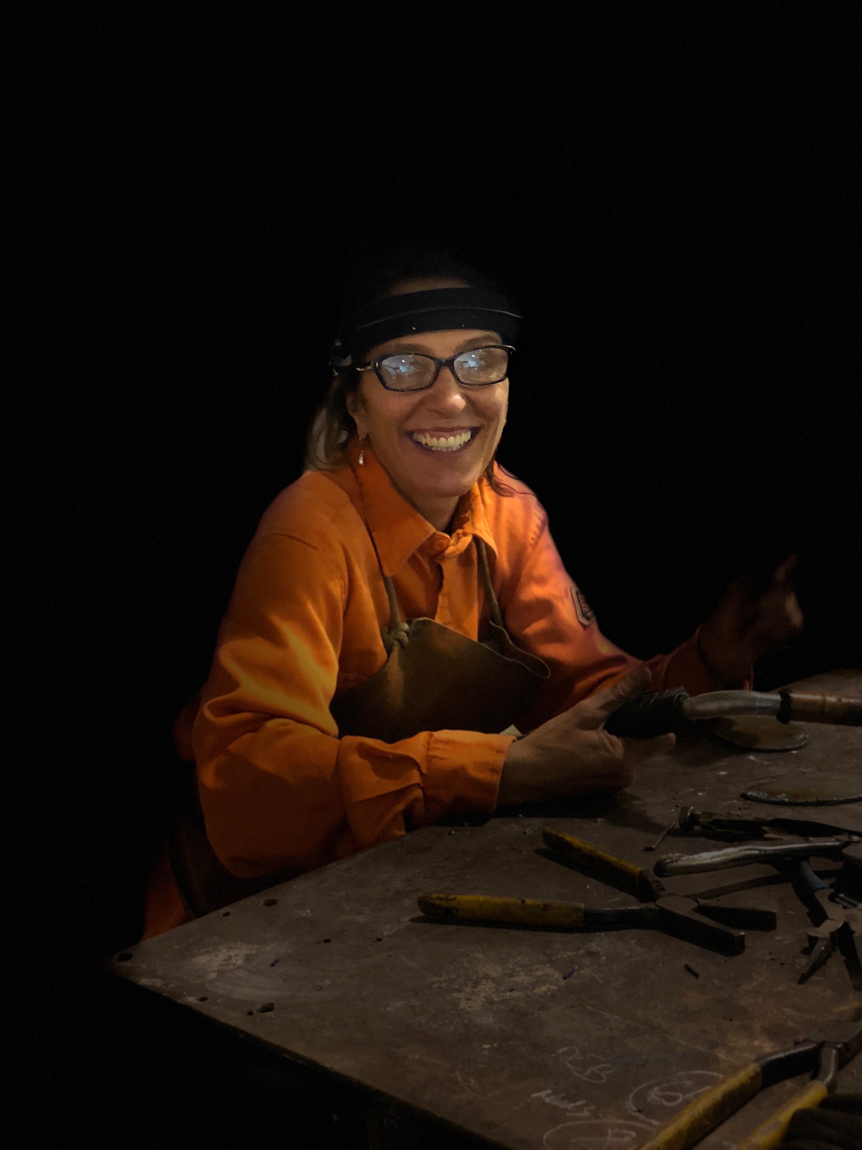 A smiling, light-skinned woman sits in a dark room. In front of her is a table with tools, including pliers. In her hand, she holds a blowtorch. She wears dark-rimmed glasses, an apron, an orange jumpsuit, and what seems to be a form of protective headgear.