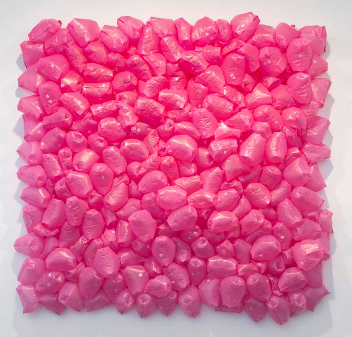A bright pink sculpture made out of tiny plastic bags filled with air. The small bags are attached to a surface in the shape of a square, making the sculpture look like a 3D painting. 