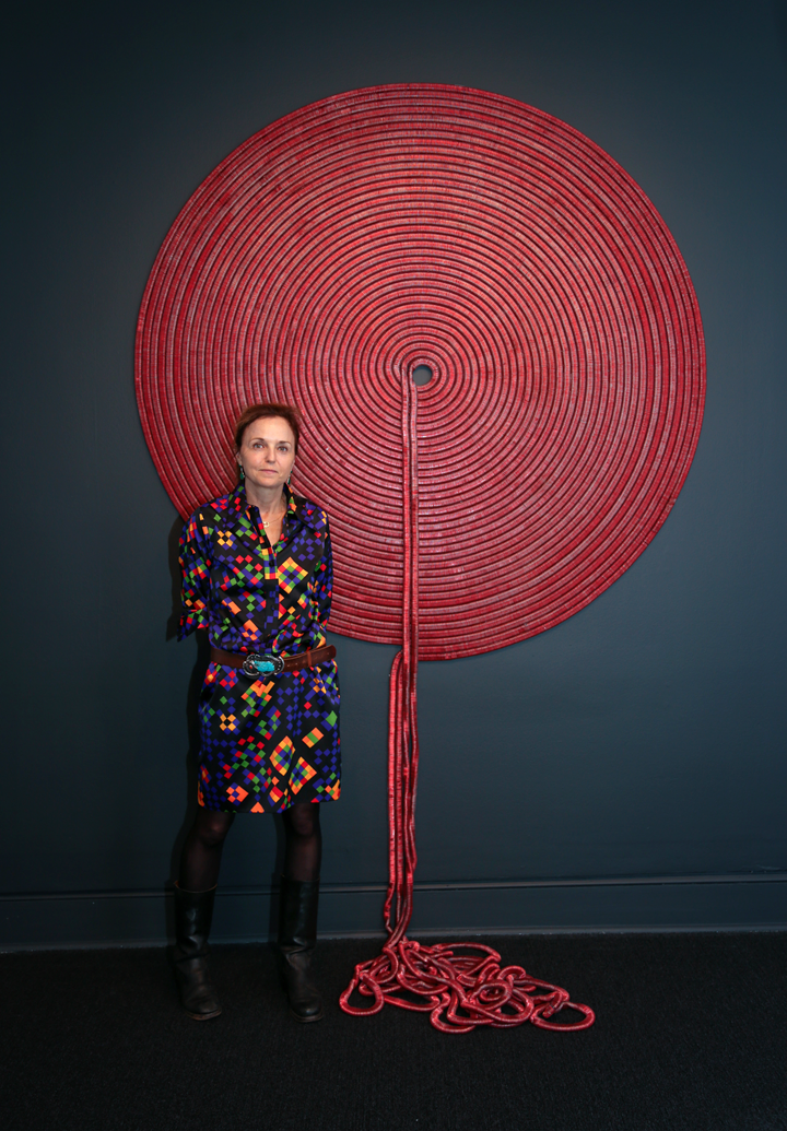 A light-skinned, red-haired woman in a colorful dress stands in front of a large, circular wall sculpture. This red-colored sculpture resembles wrapped yarn, with the loose ends spilling from the center of the sculpture to the floor.