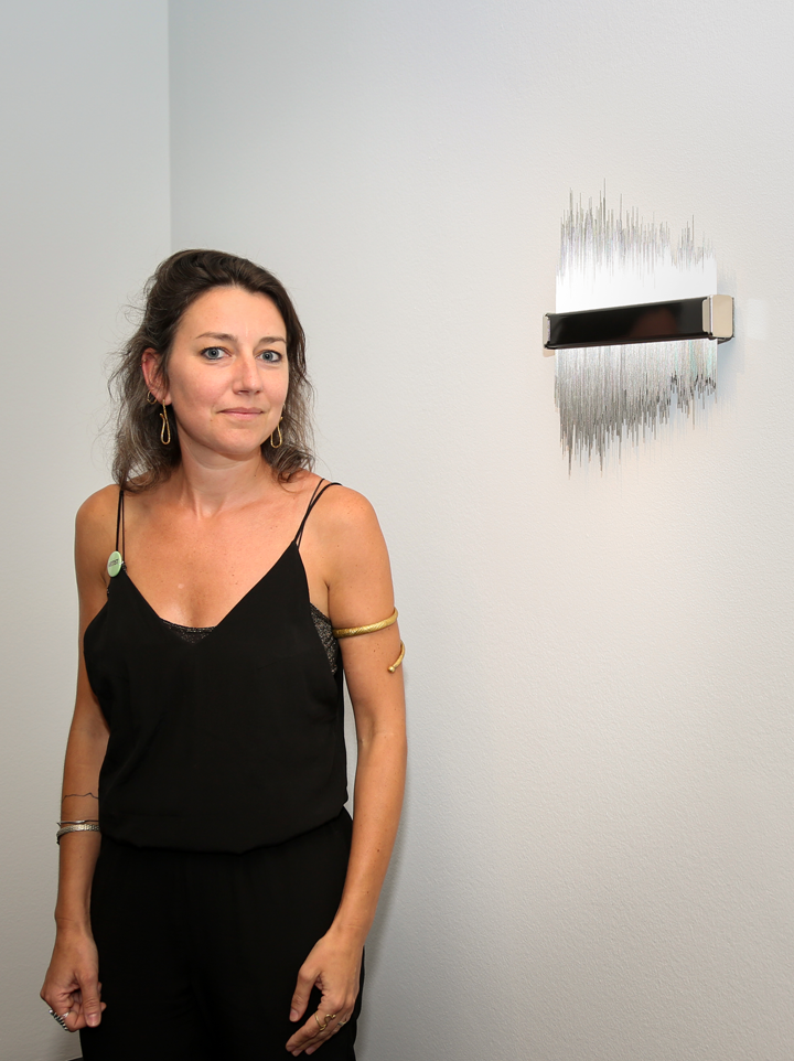 A woman with light-to-medium skin and greying brown hair stands in front of wall sculpture of metal spikes mounted with a black bar. She wears a sleeveless black dress, gold earrings, and a black bracelet around her bicep.