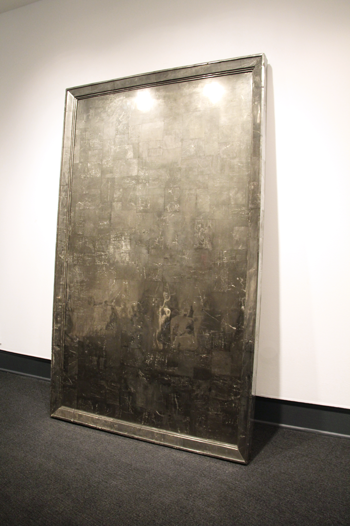A large, grey metal slab leans against a white wall. Its chromatic surface is textured  with scratches and markings.The slab has a moulded border that resembles a picture frame.