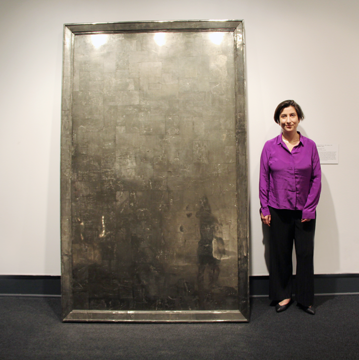 A light-skinned woman with short, dark hair stands next to a large, grey metal slab that is several feet taller than her. She wears a purple collared shirt, black trousers, and black pointed shoes.