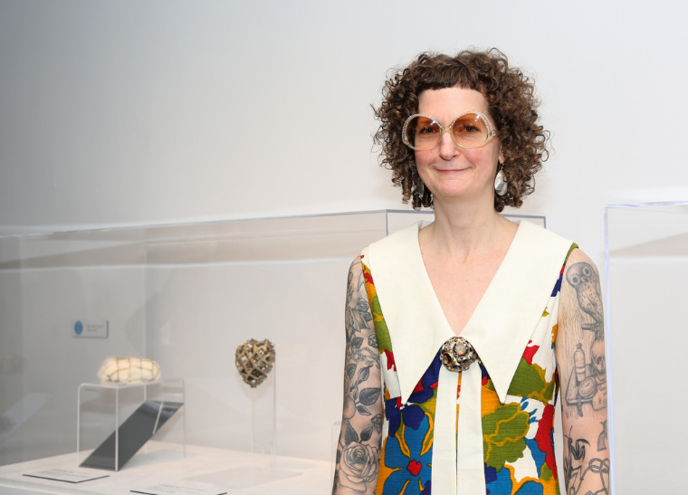 A light-skinned woman with curly brown hair stands in front of a glass display case that contains small, amorphous objects. The woman wears a colorful floral dress with a wide collar and a large metal brooch, bedazzled sunglasses, and silver earrings. Both her arms are covered in illustrative blue-ink tattoos.