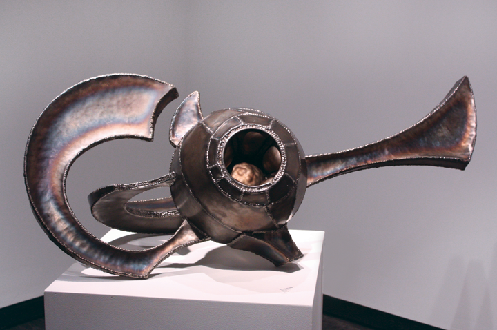 An abstract, metallic sculpture displayed on a plain white gallery pedestal in front of a grey wall. The center of the object is a hollowed-out orb. Sinuous shapes of varying sizes protrude from the exterior of the orb.