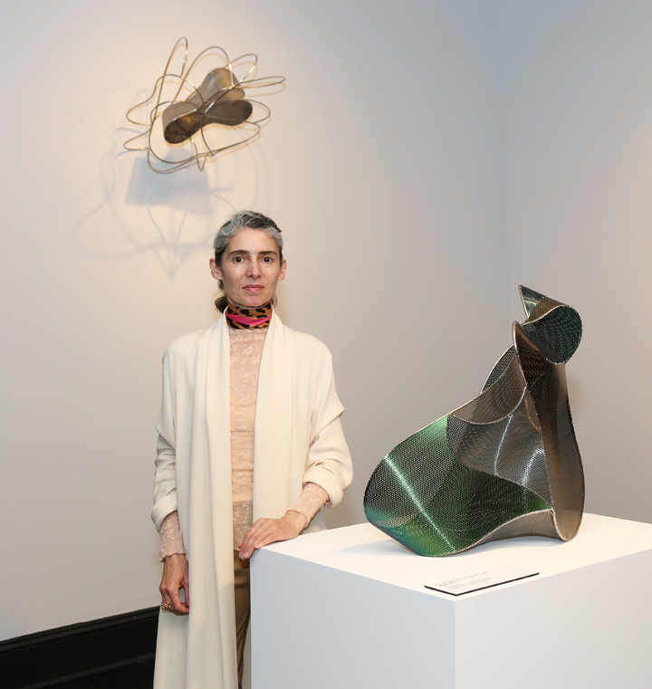 A light-skinned woman with grey hair stands beside an amorphous sculpture displayed on a white display pedestal. The woman wears a leopard-print scarf, a peach blouse, a white cardigan, and brown pants. On the wall behind her hangs a gold geometric wall sculpture.