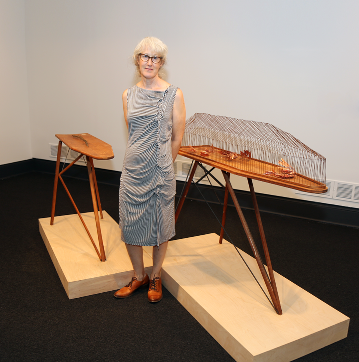 A light-skinned, light-haired woman with glasses stands in front of two sculptures, both. of which are artistic interpretations of ironing boards. One of the ironing boards has a cage-like structure on it with leaf-like sculptures encased inside. The woman wears brown shoes and a striped dress.