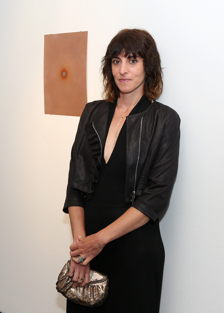 A women with brown hair and medium-light skin wearing a black leather jacket over a black dress. She carries a sequined clutch purse and stands in front of a hanging art piece. The art piece is a two-dimensional, light brown rectangle with a grey and orange circle at its center.