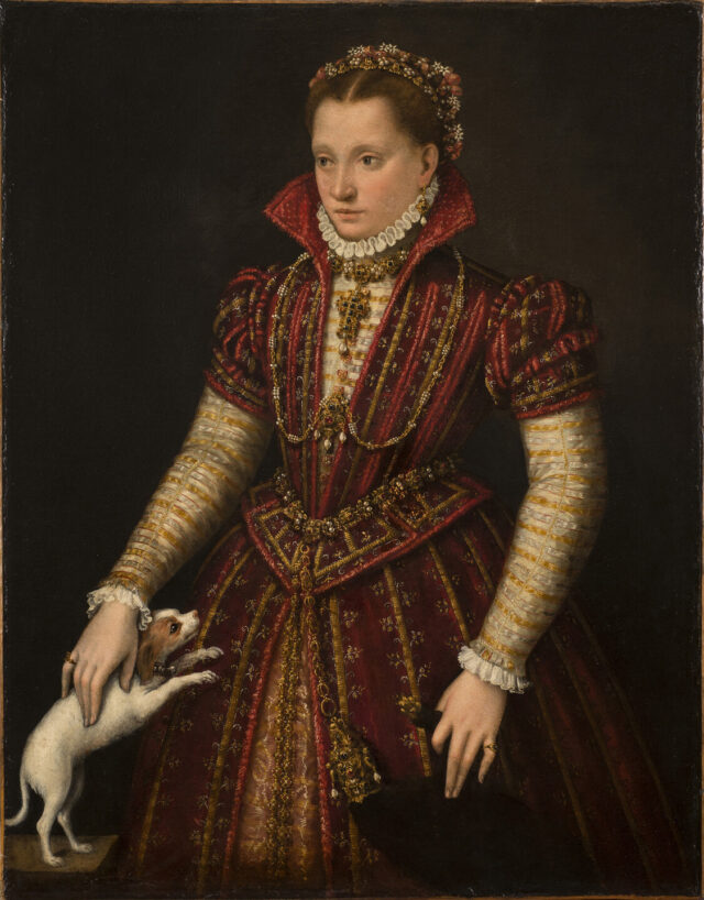 A woman with light skin tone stands in a richly brocaded red dress, her right hand reaching down to pet a small, white dog. Adorned in jewelry, she wears flowers in her hair, parted in the center. The pelt of a small mammal, its head encased in a jeweled holder, hangs from her heavily decorated belt.