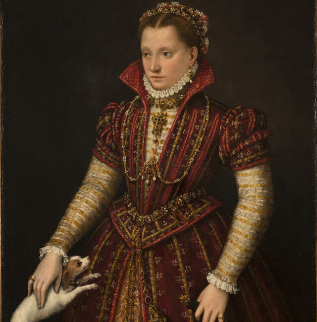 A woman with light skin tone stands in a richly brocaded red dress, her right hand reaching down to pet a small, white dog. Adorned in jewelry, she wears flowers in her hair, parted in the center. The pelt of a small mammal, its head encased in a jeweled holder, hangs from her heavily decorated belt.