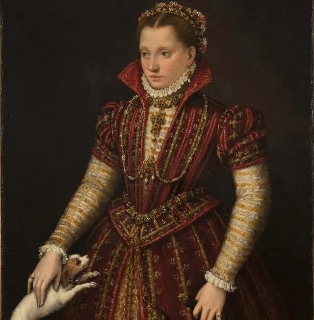 A woman with light skin tone stands in a richly brocaded red dress, her right hand reaching down to pet a small, white dog. Adorned in jewelry, she wears flowers in her hair, parted in the center. The pelt of a small mammal, its head encased in a jeweled holder, hangs from her heavily decorated belt.