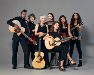 A group pf women with light, medium, and dark skin tones stand and sit before a gray wall. They are all holding a guitar and pose for the camera. 