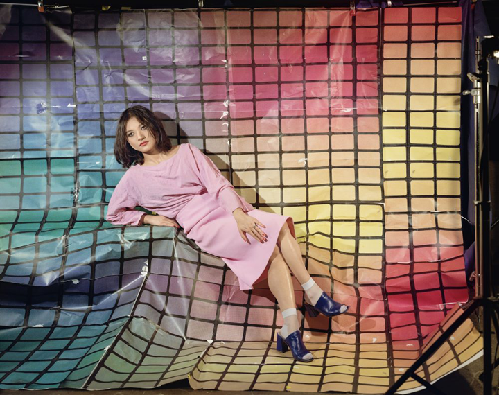 A young woman with light skin and dark hair lounges on top of multicolored, gridded fabric. The fabric conceals the object on which she sits as well as her surroundings. She wears a light pink dress, white socks, and black high-heeled shoes.