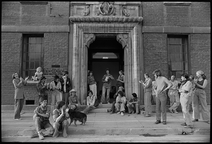 A black-and-white photograph of a group of people wearing 1970s-style fashions gathered on the sidewalk outside of a brick building. Several of the people appear to be clapping.