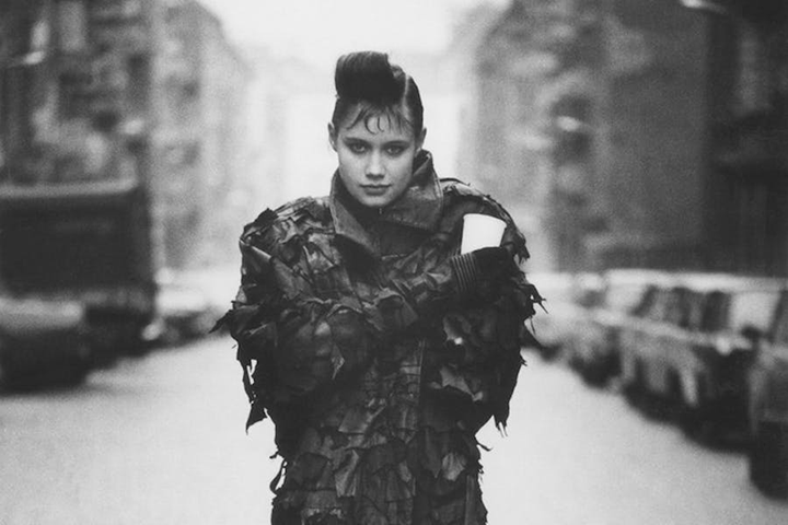 A black-and-white photograph of a fair-skinned, dark-haired woman standing in the middle of an urban street. She stares at the camera with a confident, close-lipped smile, wearing a dark jacket made from layers of tattered fabric. She holds a white cup in her gloved hand.