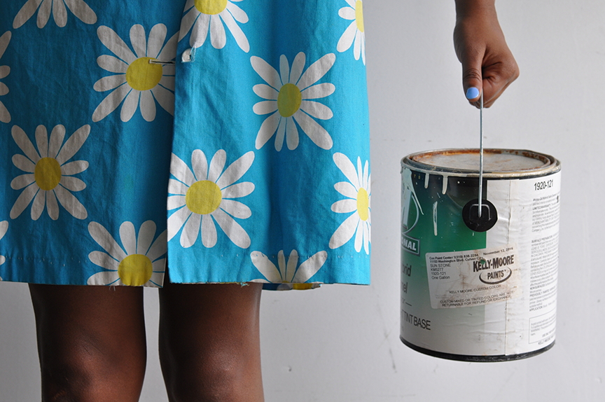 A dark-skinned woman wears a blue skirt with white daisies printed on it. She holds a can of paint in her left hand. The photo is cropped so that only her knees, skirt, hand, and paint can are visible. She has blue nailpolish.