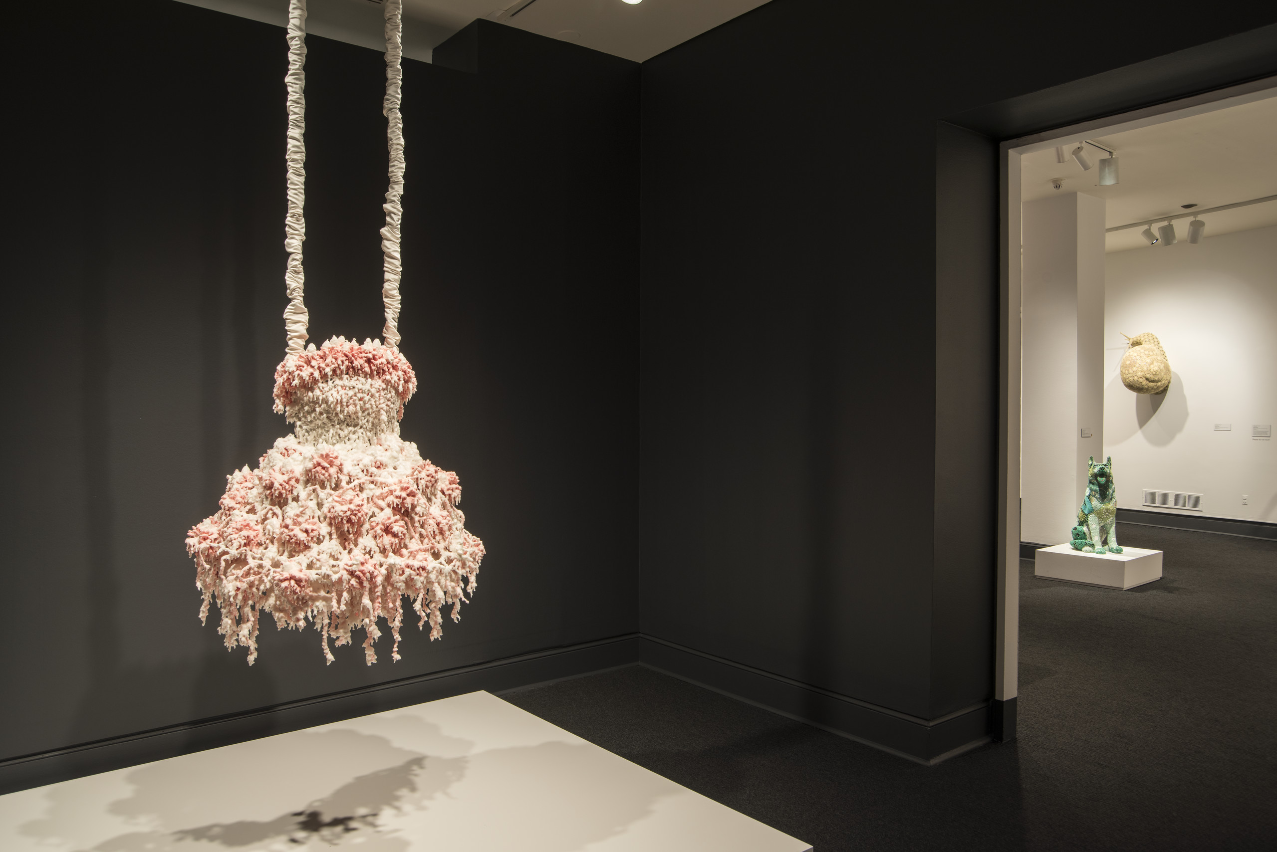A sculpture hangs in a dark gallery. The sculpture comprises layers of melted pink and white wax that form a dress-like shape hanging from satin-wrapped chains. Its color, shape, and bumpy, lacy texture, evoke a frilly tutu, lavishly frosted wedding cake, or coral.