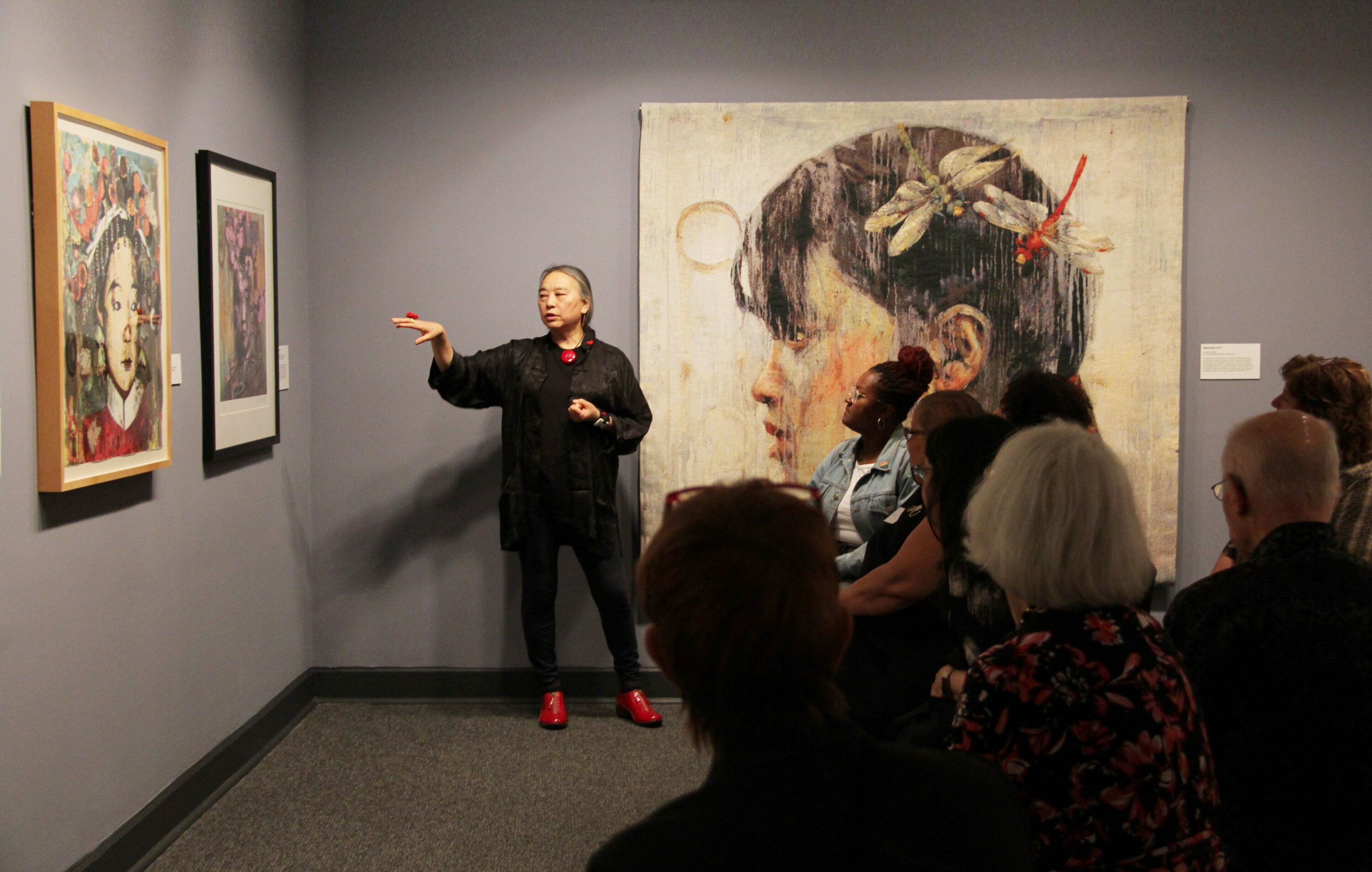 Hung Liu speaks to an audience about her paintings of Chinese women, which hang on the walls around her. Liu is a light-skinned adult woman with gray hair in a low ponytail, dressed in black with red jewelry and shoes. She stands, gesturing with one hand at a painting to her right.