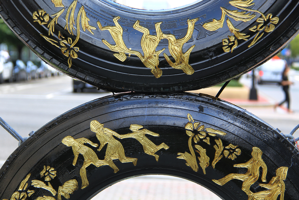 An image of black car tires mounted on top of one another. The image only shows the bottom-half of the top tire and the top-half of the bottom tire. Yellow-beige illustrations flowers and running figures are carved into the rubber of the tires. The running figures are silhouettes of a man, a woman, and a child.