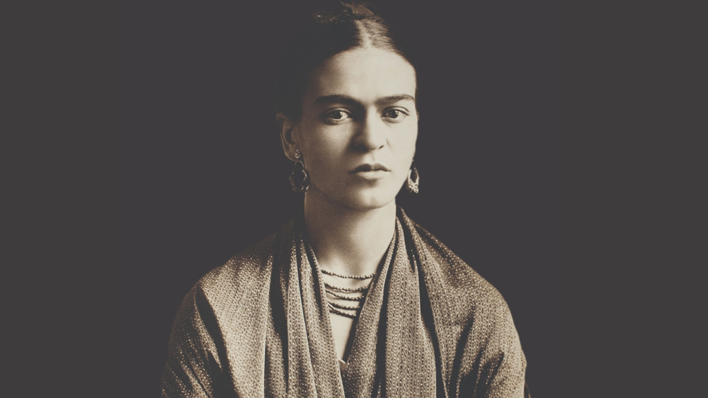 A sepia portrait of Frida Kahlo from the chest-up. Kahlo wears a shawl, five beaded necklaces, and pear-shaped drop earrings. Her dark hair is pulled back into a bun.