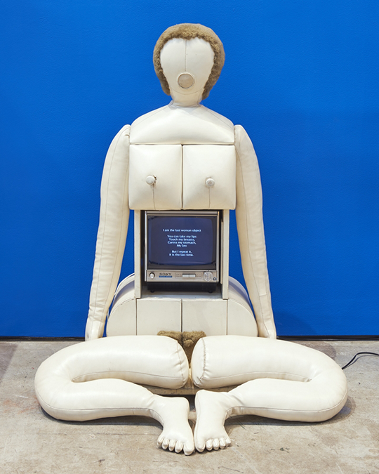 An abstract sculpture of a woman against a cobalt blue background. The sculpture's limbs are made of stuffed white fabric while its torso is made of an old-fashioned Sony television. There is white text on the screen. The figure has no face and its legs are detached from its body.