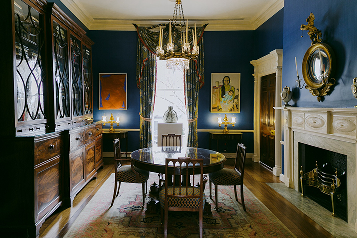 A dining room interior with a circular dining table and four chairs standing the center on a faded rug. The walls are painted dark blue. There is a window located directly across the room from where the photo is taken. Two paintings hang on either side of the window. The room includes a white fireplace to the left the dining table and chairs. A circular mirror hangs above the fireplace. To the right of the table and chairs is a large wooden cabinet with glass doors that takes up the entire wall.